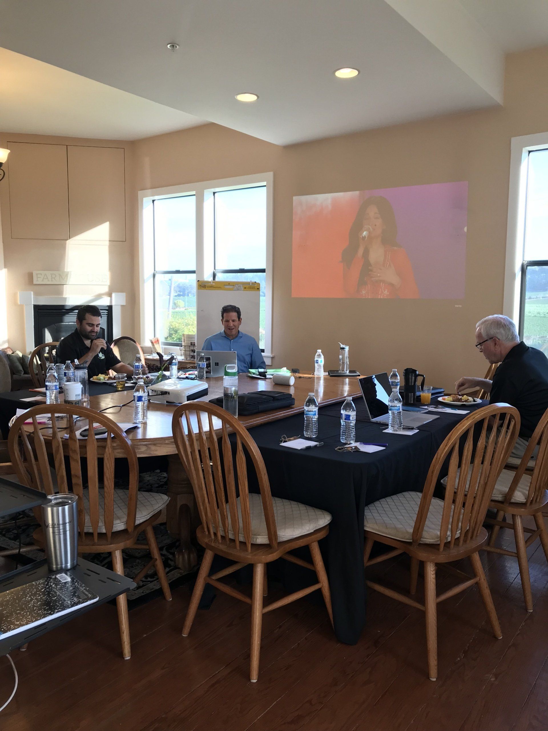 A group of people are sitting at tables in a room with a projector screen on the wall.