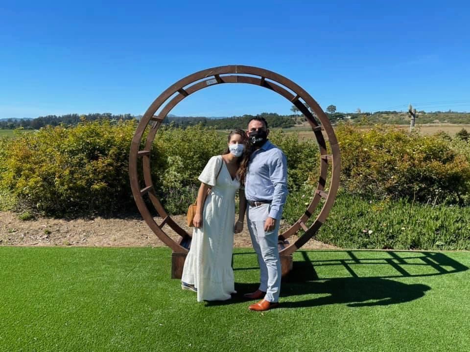 A bride and groom wearing face masks are posing for a picture in front of a wooden arch.