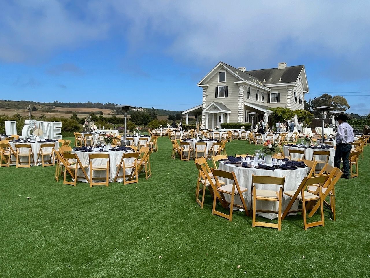 Tables and chairs are set up in a grassy field in front of a large house.