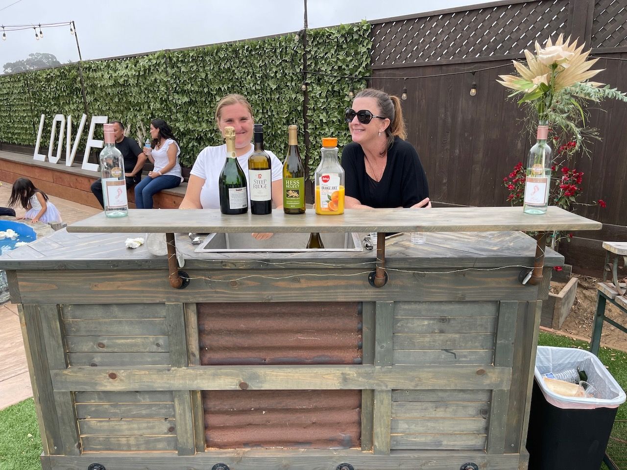 Two women are standing behind a wooden bar with bottles of wine on it.