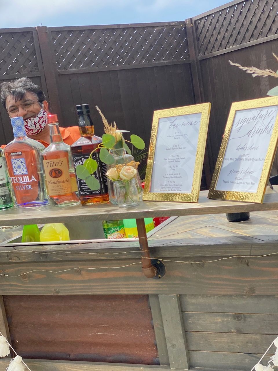A man wearing a mask is standing behind a wooden bar filled with bottles of alcohol.