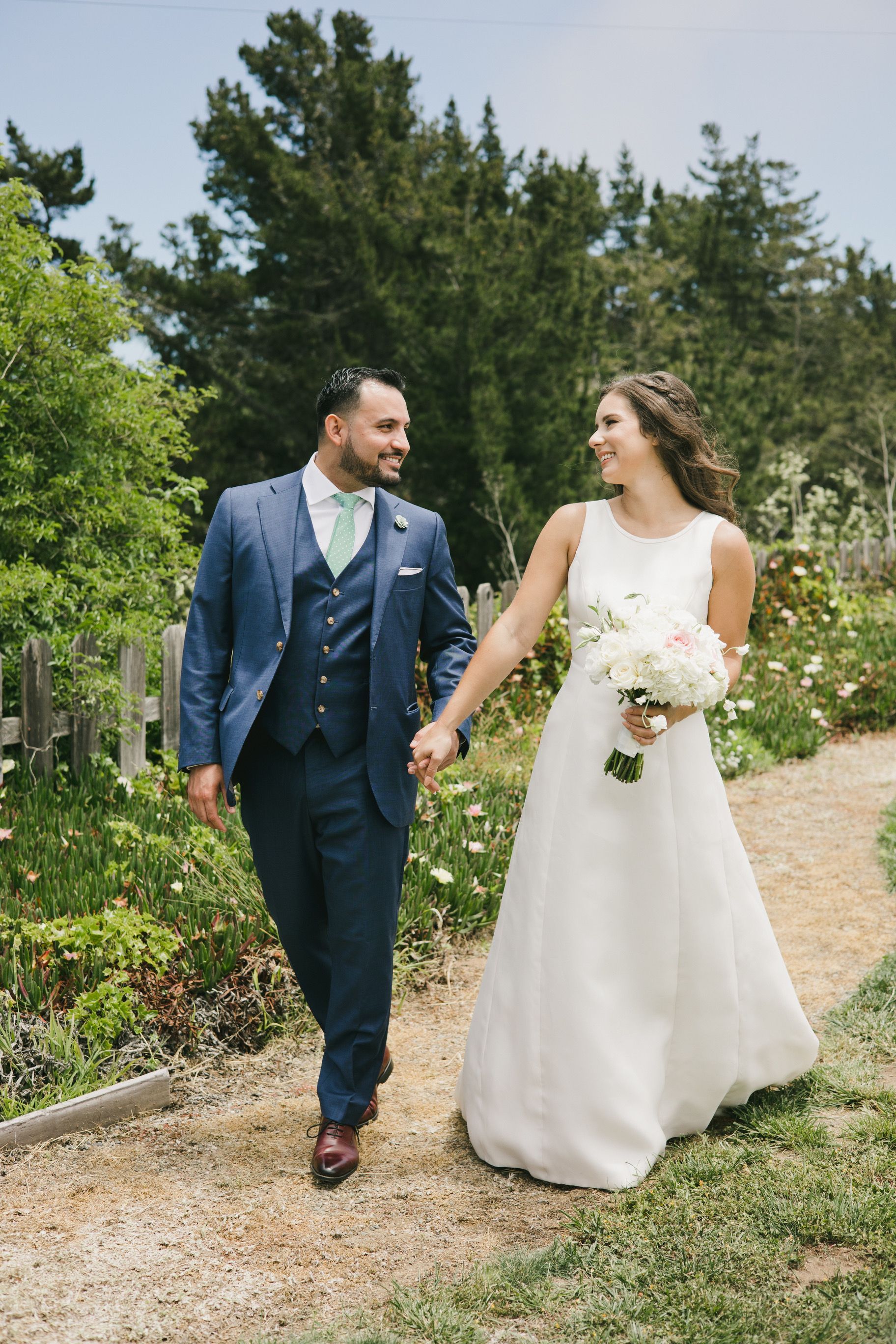 A bride and groom are walking down a path holding hands.