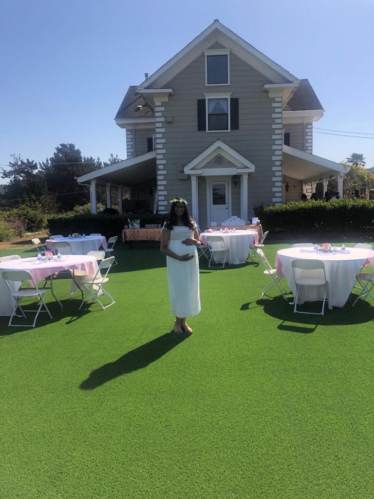 A pregnant woman is standing in front of a large house with tables and chairs set up for a baby shower.