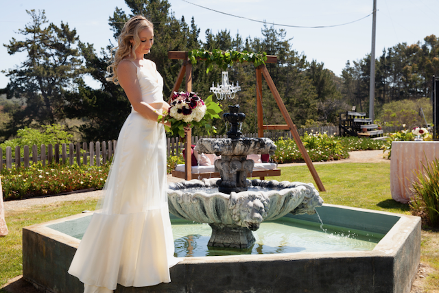 A bride in a wedding dress is standing in front of a fountain holding a bouquet of flowers.