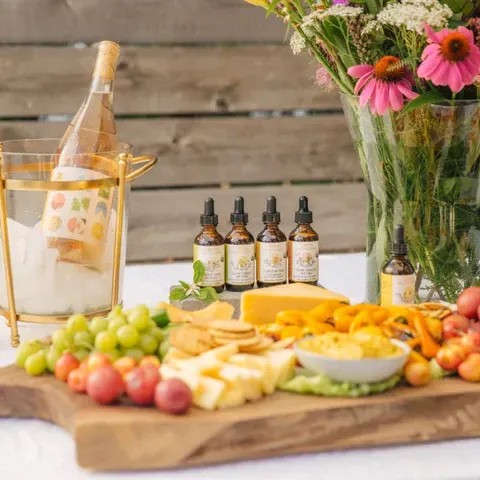 Charcuterie board with fruit, cheese, crackers, and essential oils. A bottle of wine chills in an ice bucket, flowers in a vase.