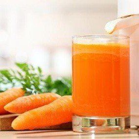 Carrot juice being poured from a juicer into a glass, with carrots and greens in background.