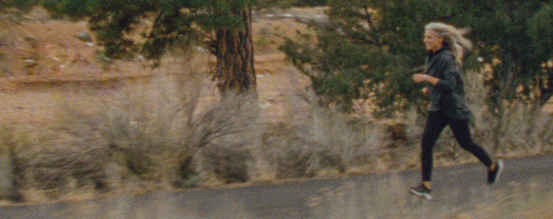 A woman with blonde hair runs on a path in a natural setting. She wears dark athletic clothing.