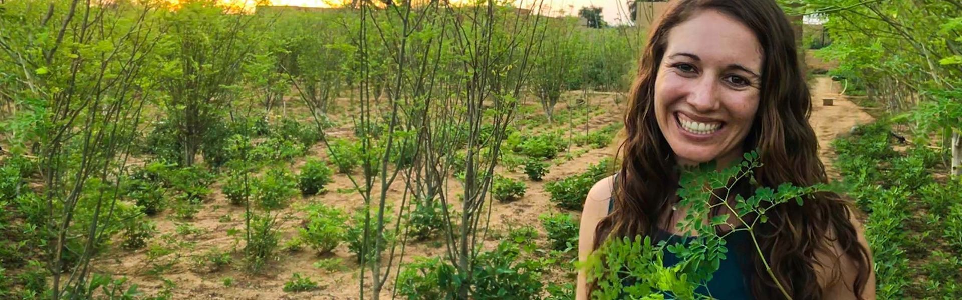 A smiling woman surrounded by greenery in a field, possibly a farm or garden.