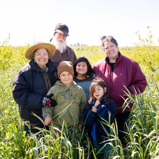 Family standing in a field of tall grass, smiling at the camera. Adults and children, diverse skin tones, sunny day.