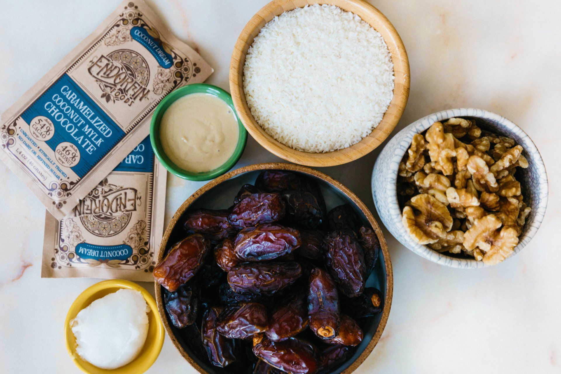 Ingredients for a recipe: dates, walnuts, shredded coconut, chocolate, tahini, and coconut oil in various bowls.