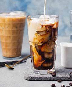 Iced coffee being poured into a glass with ice, creating a swirl of cream and coffee. A second glass of iced coffee and coffee beans are also visible.