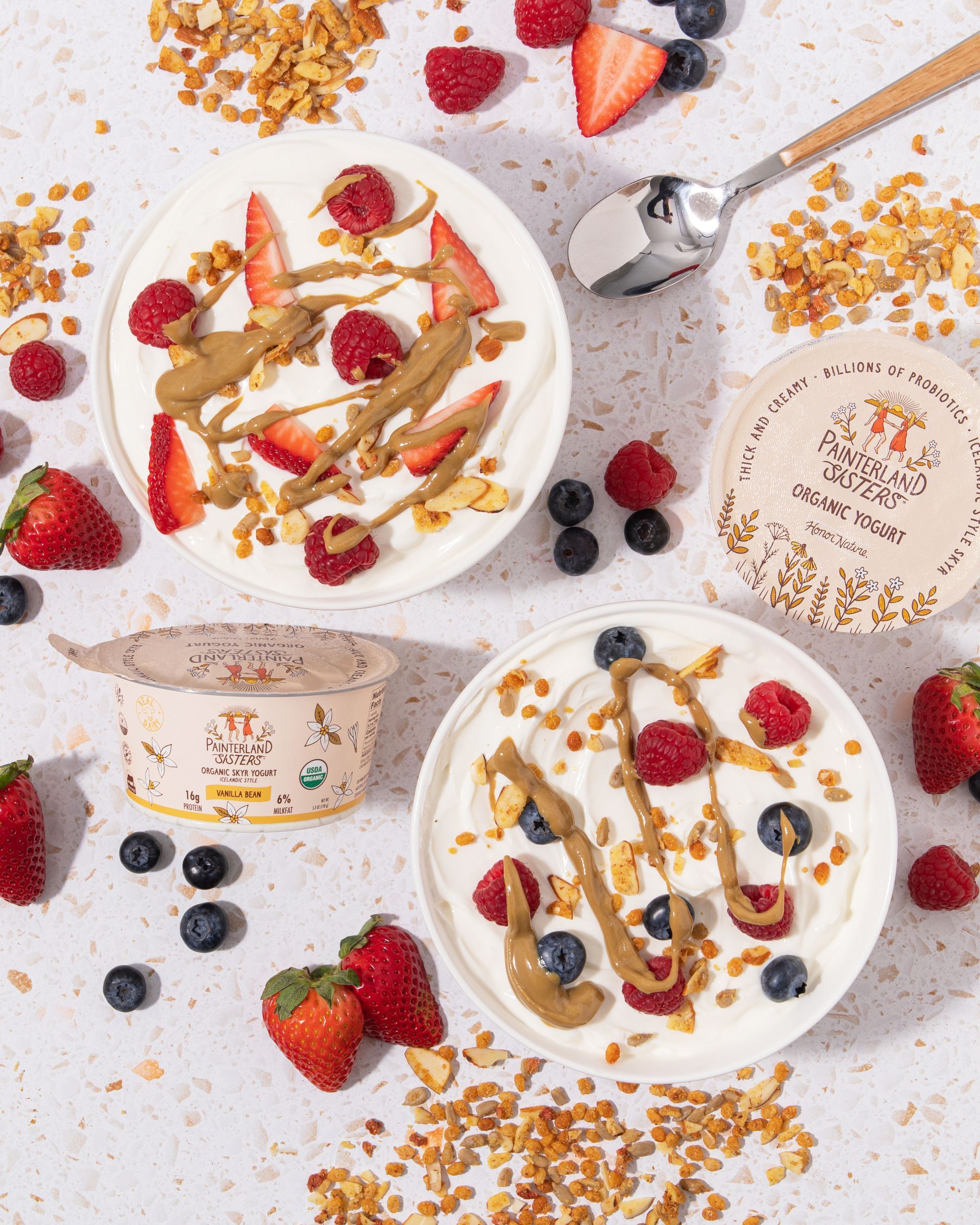 Two bowls of yogurt topped with berries, nuts, and almond butter, next to two yogurt containers and a spoon.