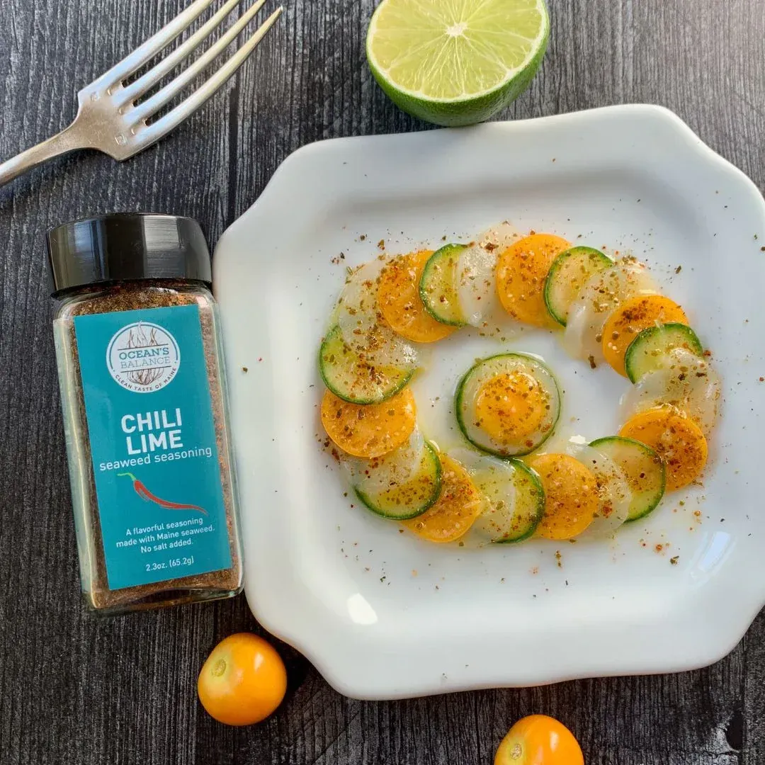 A plate of sliced vegetables (yellow and green) arranged in a circle, seasoned with chili lime spice, lime and golden berries on a dark wooden surface.