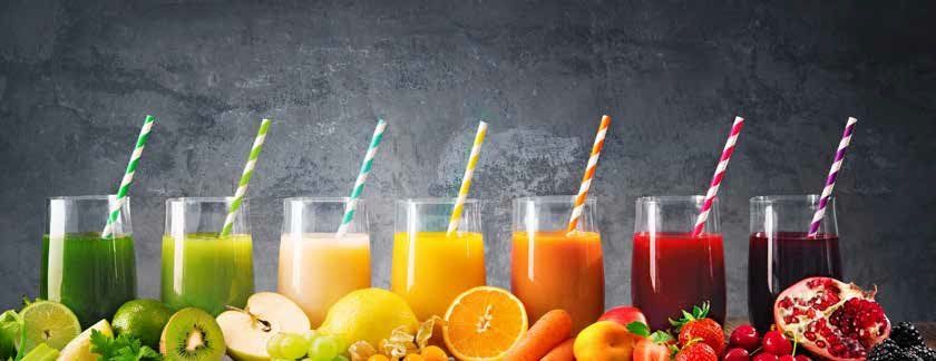 A row of colorful juices with straws, next to the fruits they are made from, against a dark background.