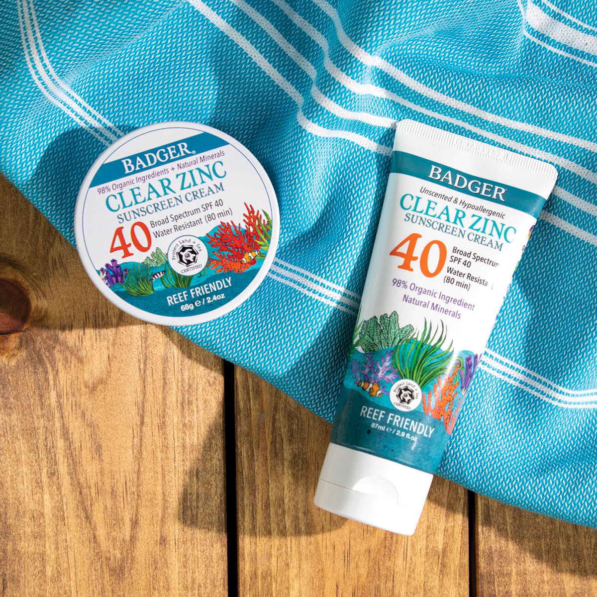 Two Badger Clear Zinc Sunscreen products (tub and tube) on a blue towel, on a wooden surface, with a beach theme.