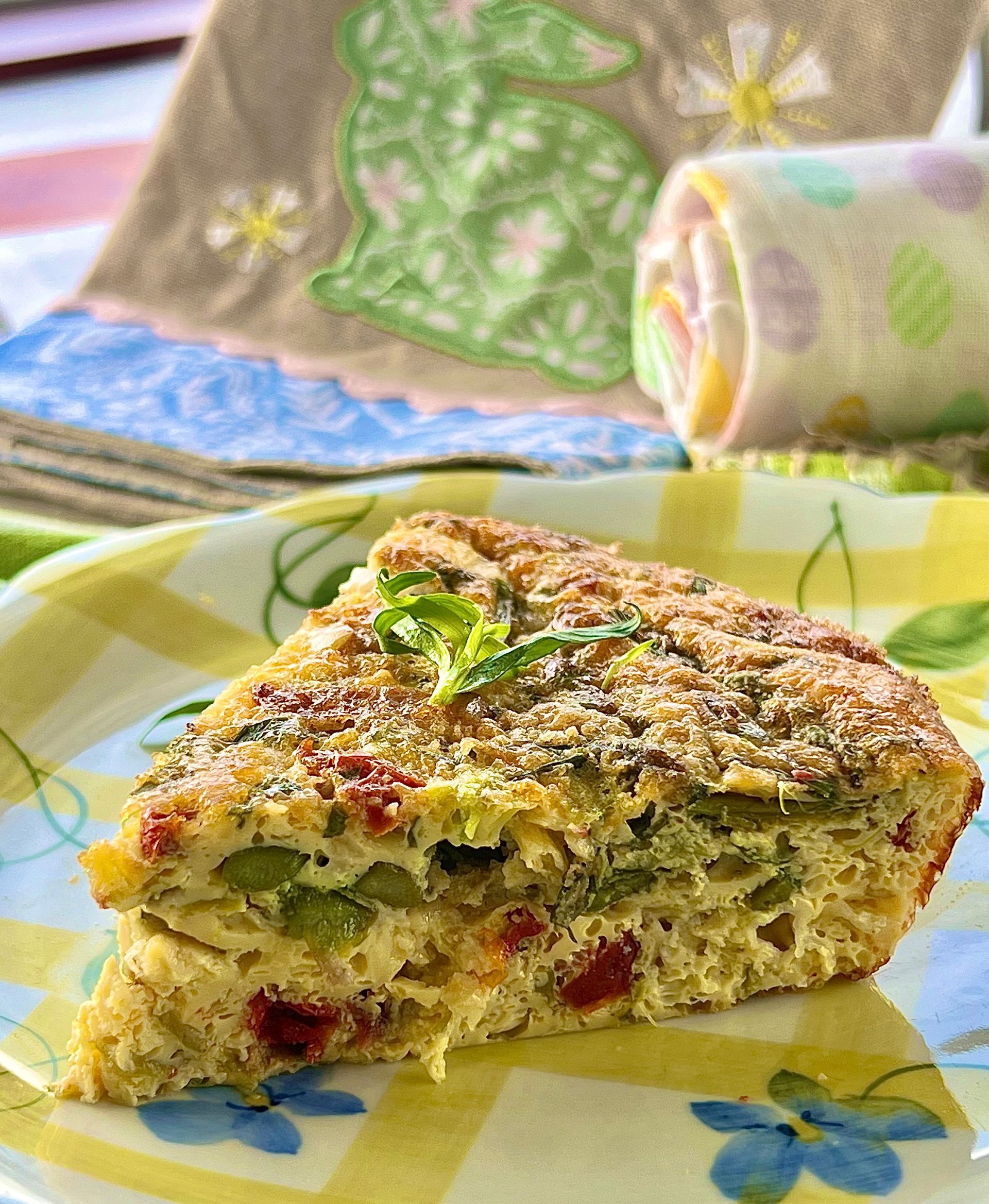Slice of quiche on a floral plate, featuring asparagus and red tomatoes. The background has a rolled pastel towel and a bunny-themed cloth.