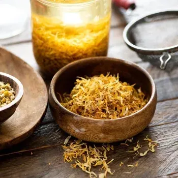 Dried herbs in wooden bowls and a jar, with a strainer on a wooden table.