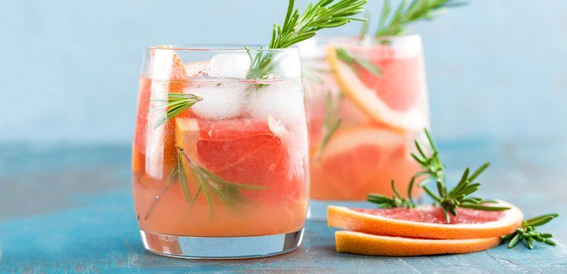 Two glasses of pink grapefruit cocktails garnished with rosemary and grapefruit slices on a blue wooden table.