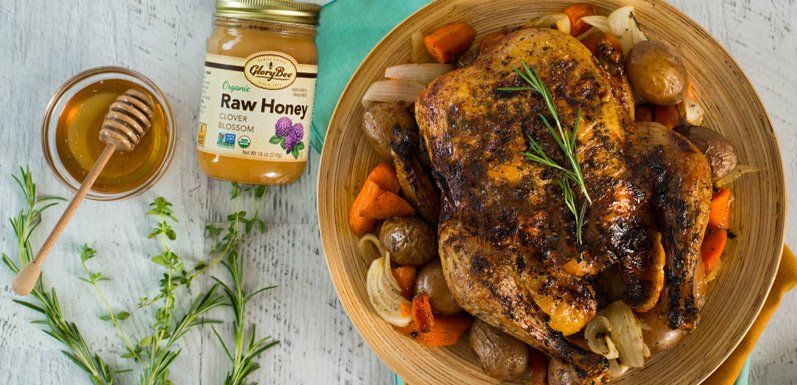 Roasted chicken and vegetables in a wooden bowl next to honey with a honey dipper and sprigs of rosemary.