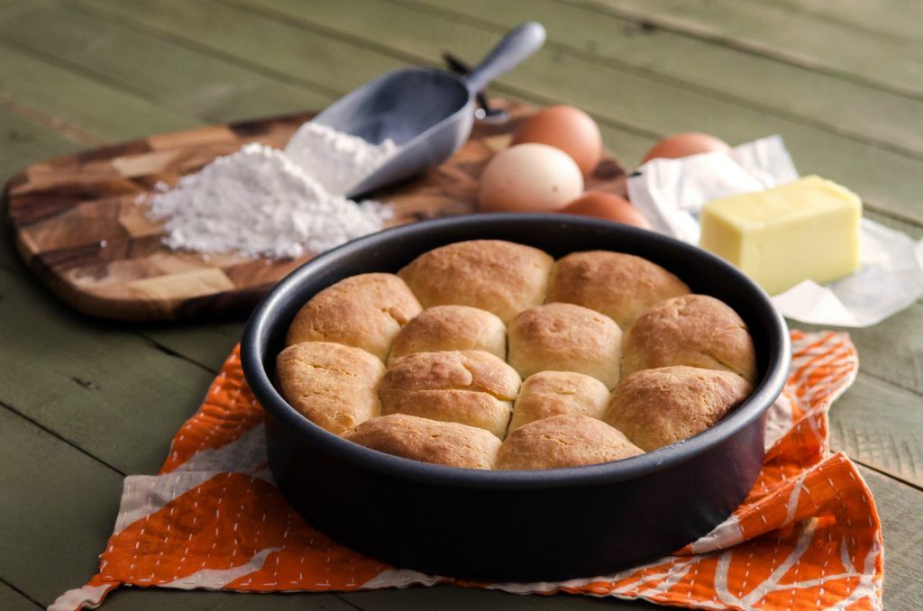 Baked bread rolls in a round baking pan, ingredients like flour, eggs, and butter in the background.