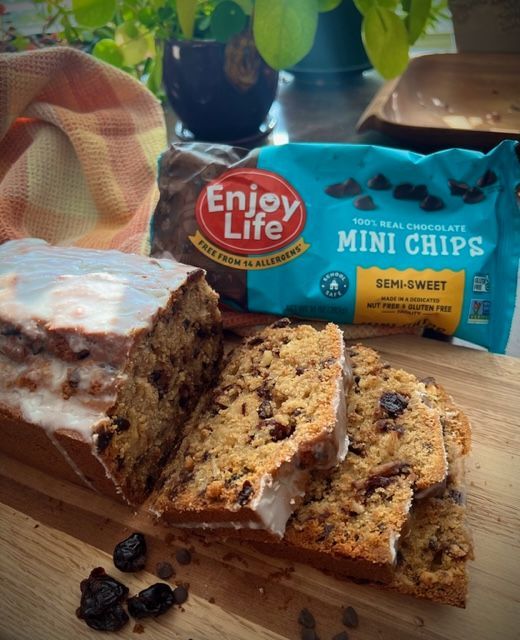 A loaf of cake with chocolate chips and glaze, next to a package of Enjoy Life mini chocolate chips, on a wooden board.