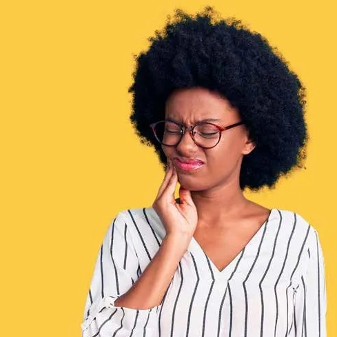 Woman with glasses, dark curly hair, and a striped shirt clutches her jaw, showing a pained expression on a yellow background.