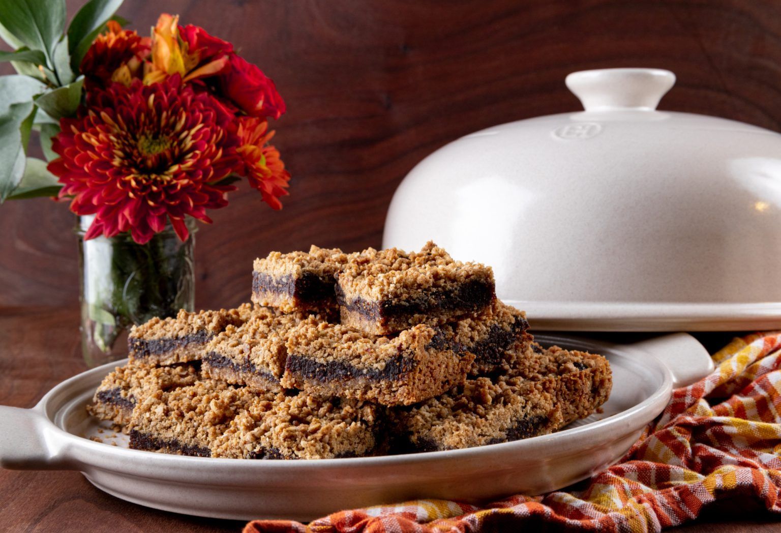 Brownies with crumb topping on a white platter, near flowers and a covered dish.