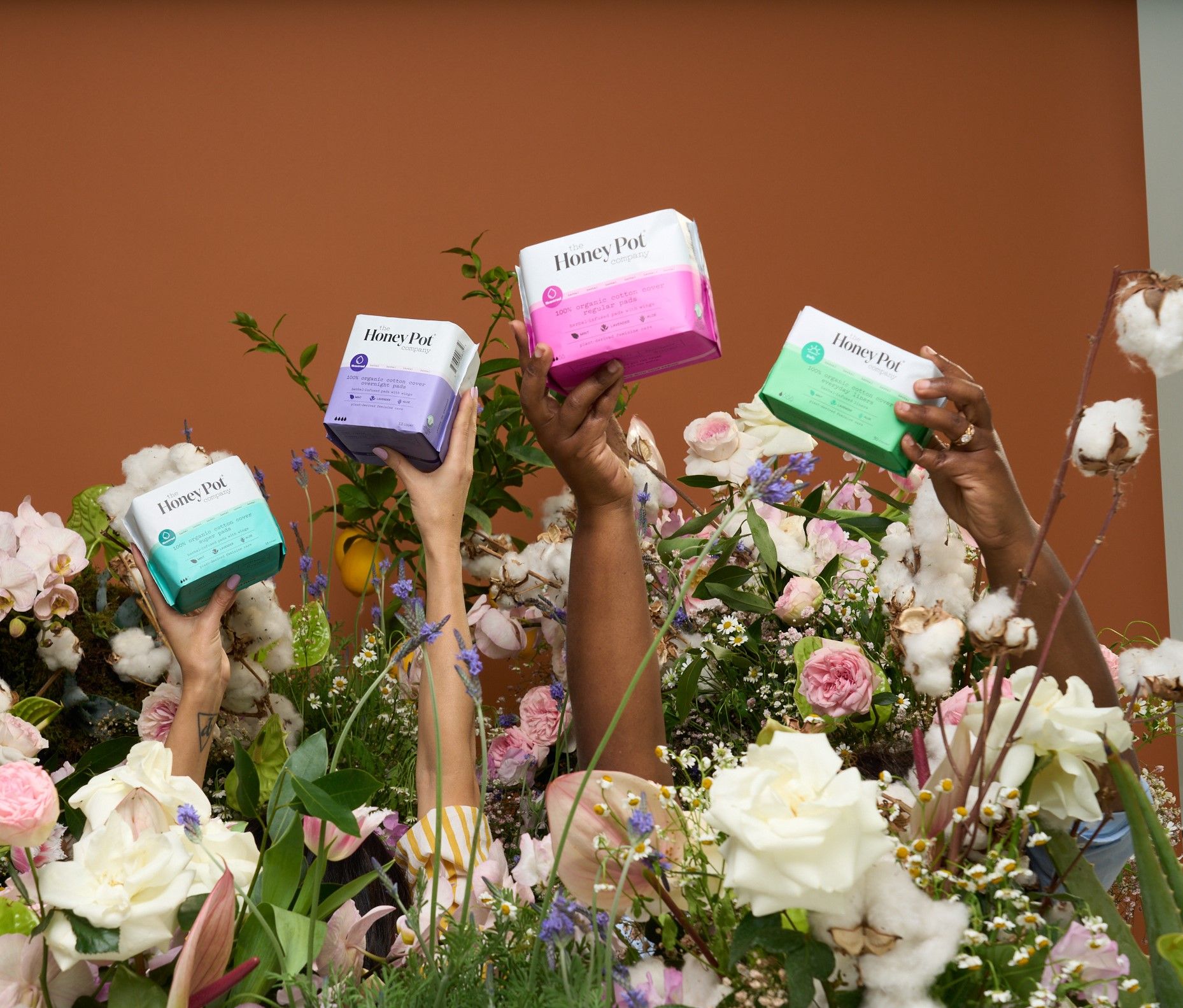 Four hands holding colorful boxes of period products, surrounded by flowers against a brown background.