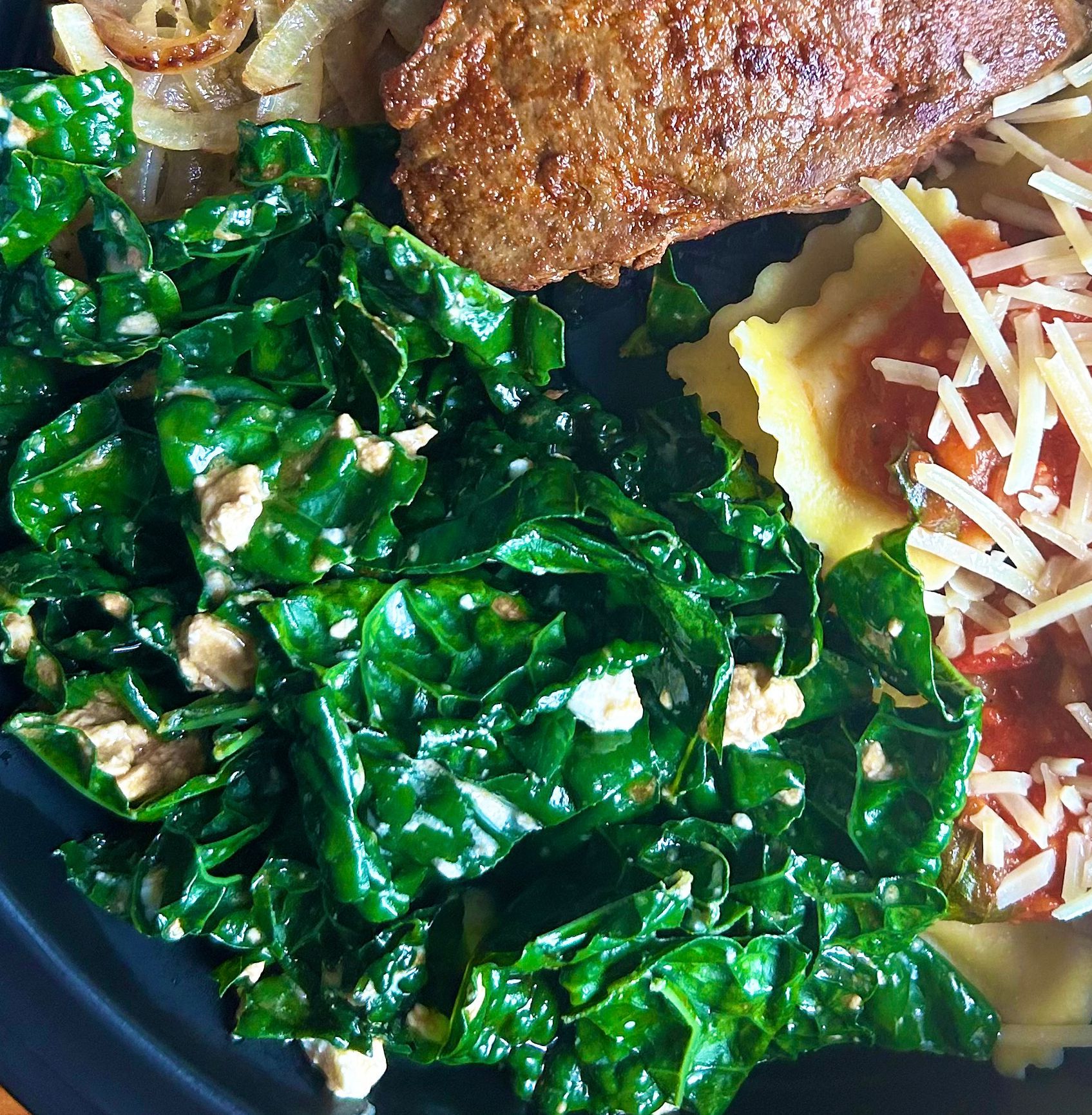 A plate with sauteed kale, a piece of meat, ravioli with red sauce and cheese, and some sauteed onions.