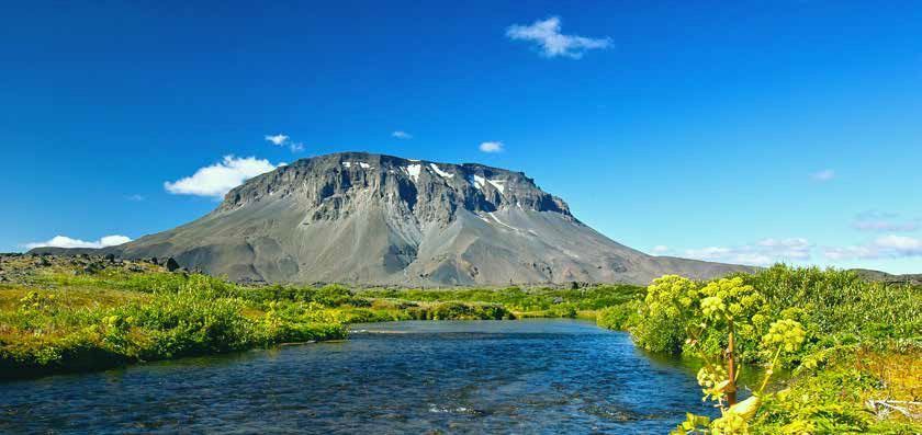 A mountain with patches of snow sits under a bright blue sky. Green vegetation lines a body of water.
