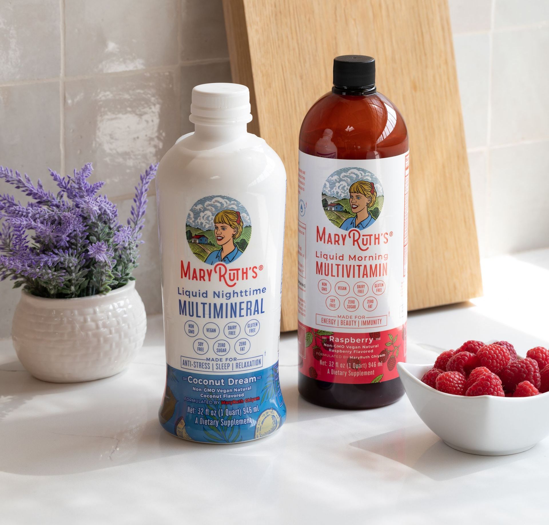 Two bottles of Mary Ruth's liquid multivitamin and multimineral, plus a bowl of raspberries and a lavender plant, on a counter.
