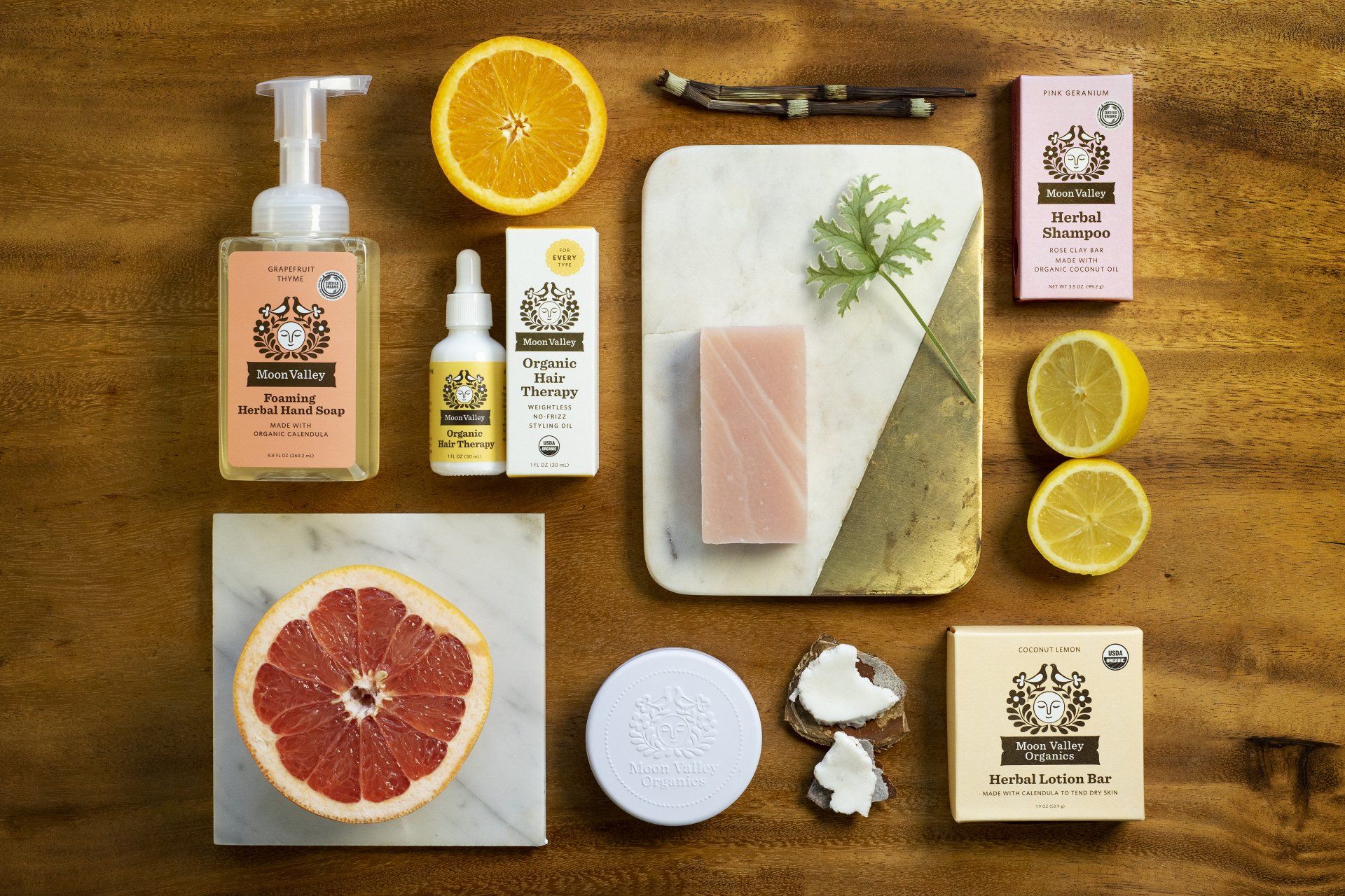 Flat lay of beauty products, including soap, serum, and citrus fruit on a wooden surface. Pink, yellow, and white items are arranged around the fruit.