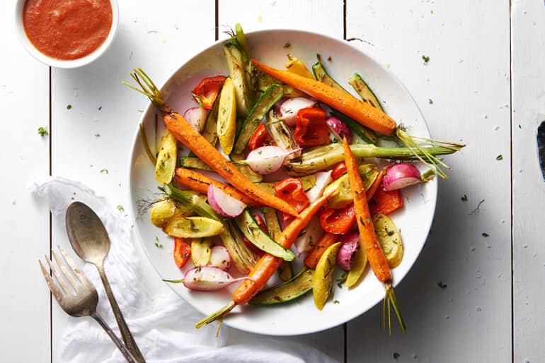 Roasted vegetables (carrots, zucchini, radishes, onions) in a white bowl, served with tomato sauce and silverware on a white surface.
