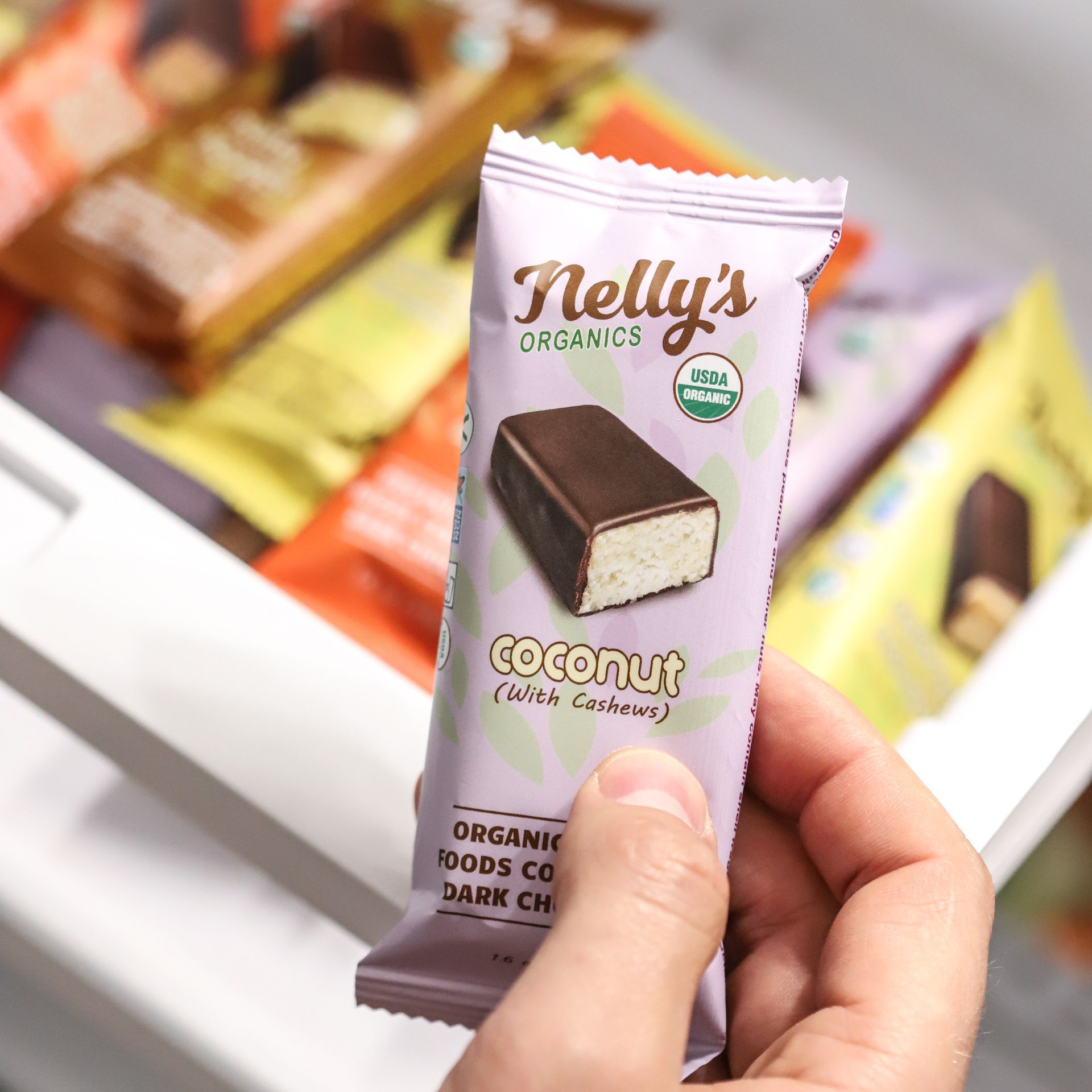 Hand holding a purple Nelly's Organics Coconut snack bar, with other colorful bars in the background.