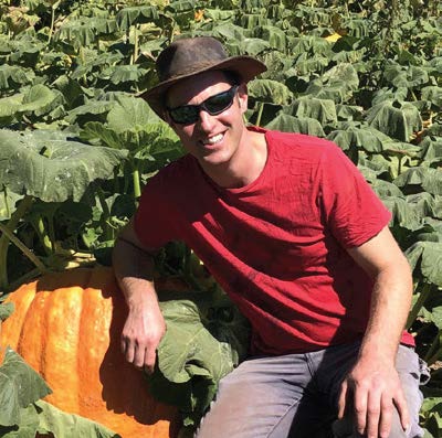 Man in red shirt and hat smiles, leaning on a large orange pumpkin in a pumpkin patch.