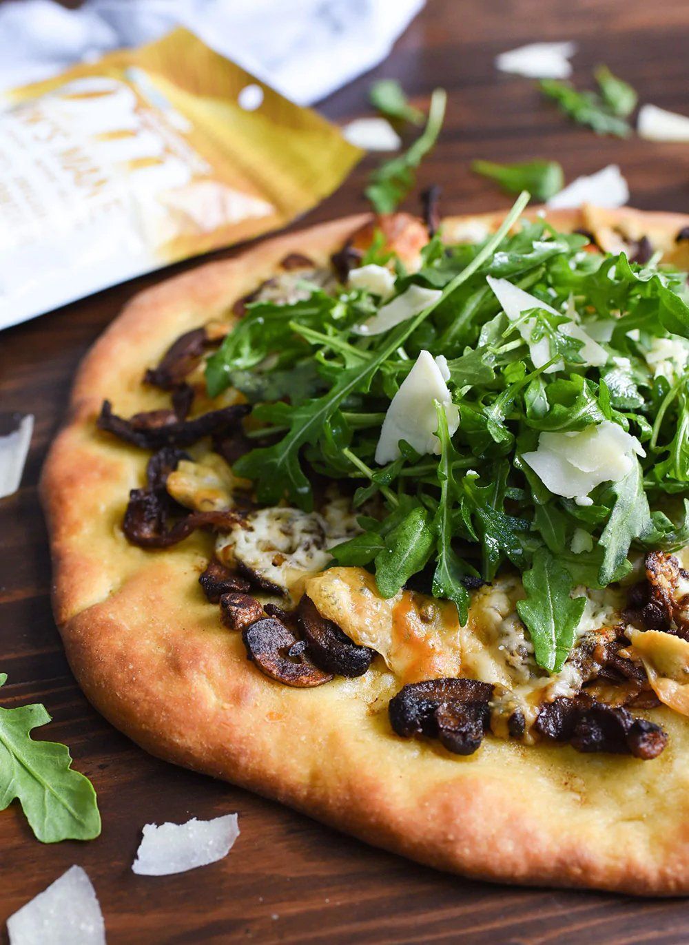 Flatbread pizza topped with mushrooms, arugula, and shaved parmesan cheese on a wooden table.