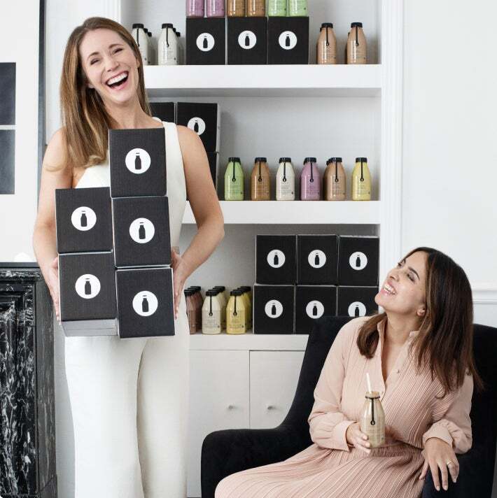 Two women laughing, one holding boxes of product, the other seated with a beverage. The setting is a bright, modern space with shelves of products in the background.