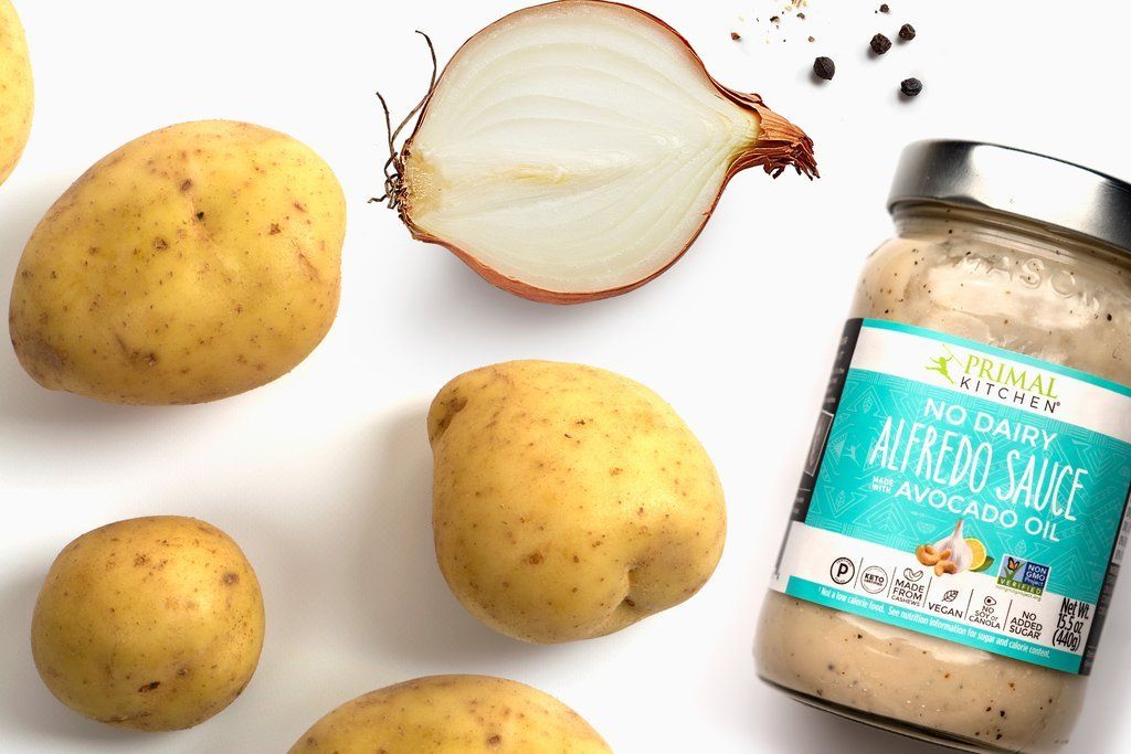 Potatoes, half an onion, and a jar of dairy-free Alfredo sauce on a white surface.