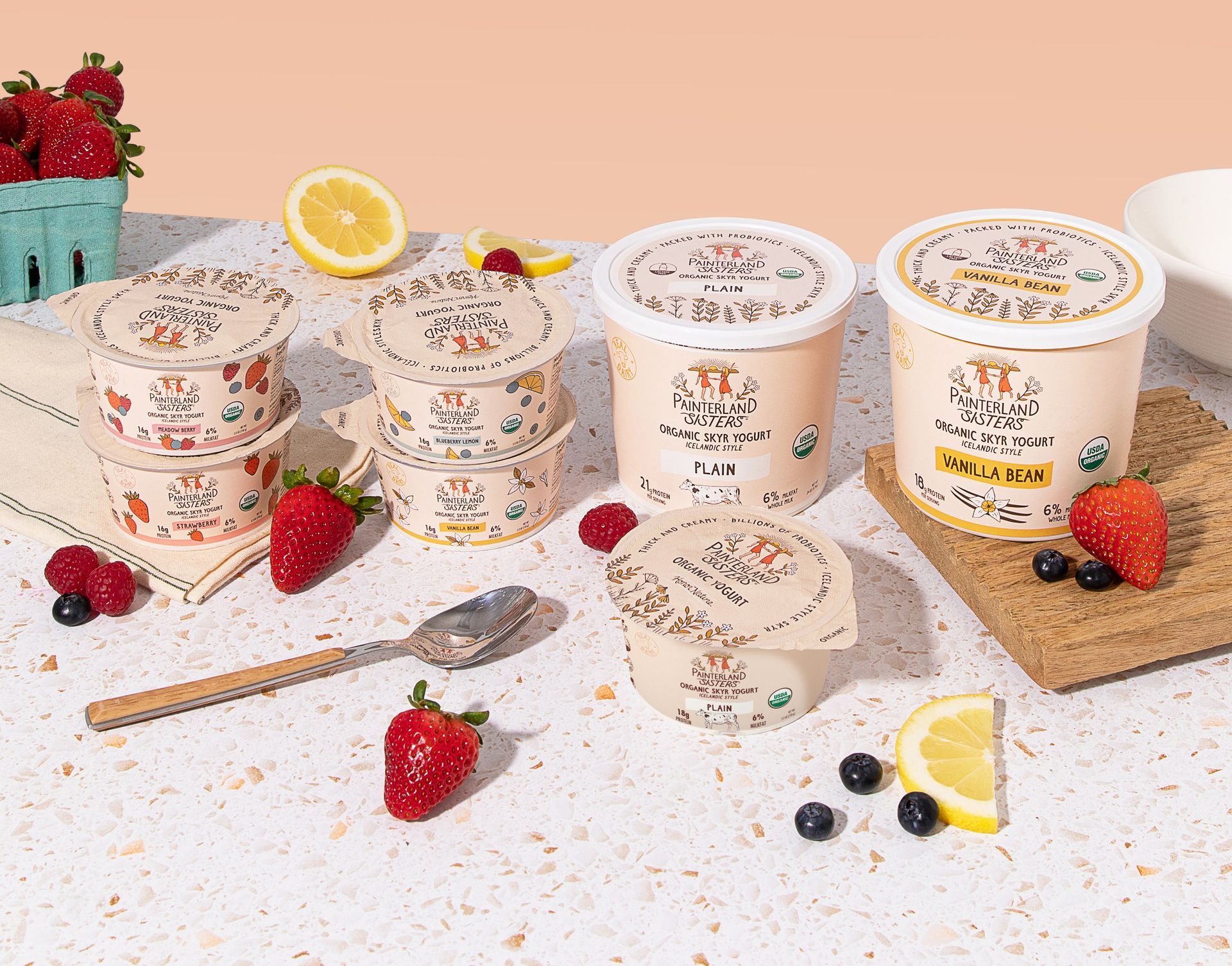 Yogurt cups with fruit like strawberries, raspberries, and blueberries are arranged on a table with a spoon and a lemon half.