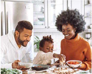 A Black family of three cooking together in their kitchen. The father and mother are smiling, and the child is helping.
