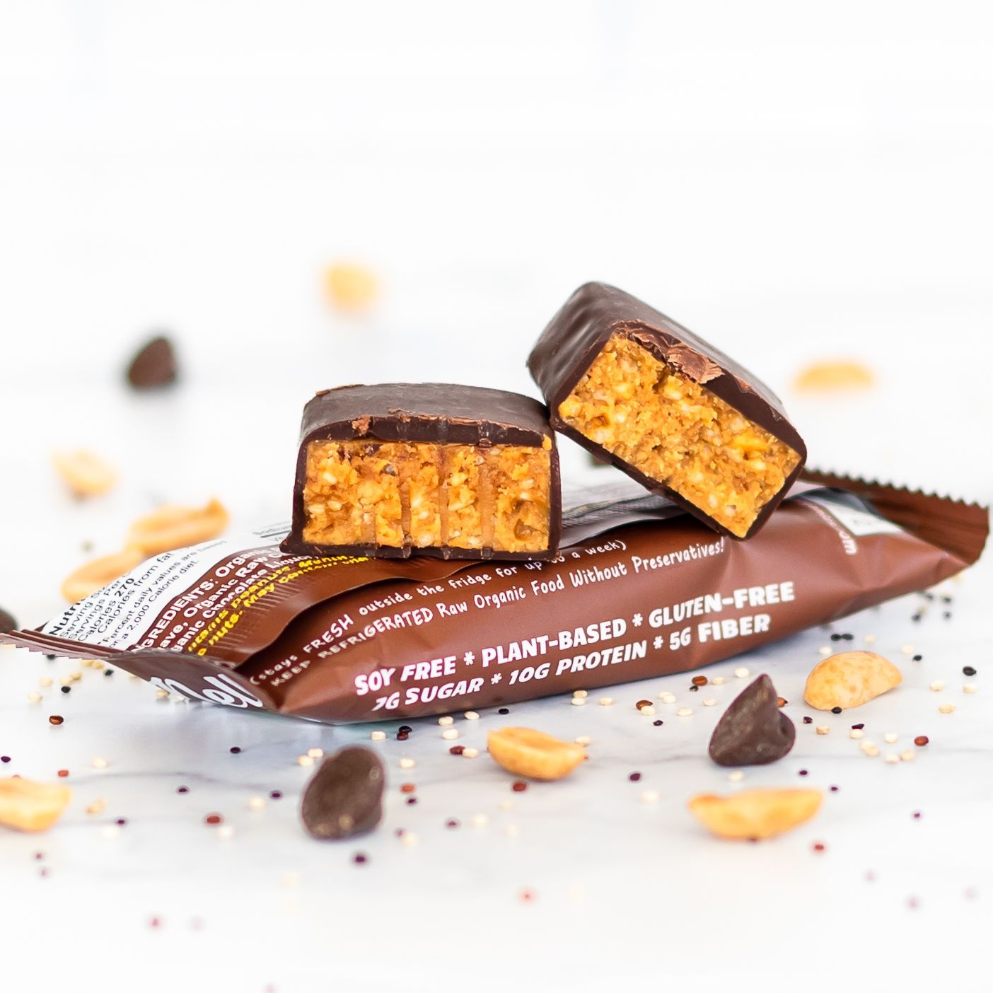 Chocolate-covered peanut butter snack bar cut in half, resting on its wrapper. Peanuts and chocolate chips are scattered around.