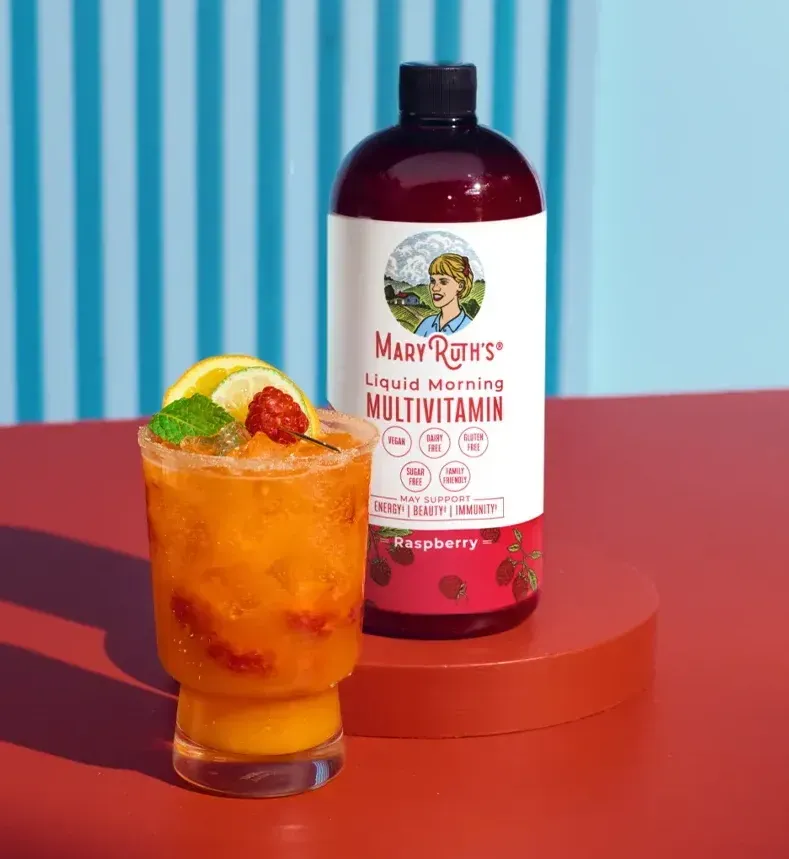A glass of raspberry-colored cocktail with garnish and a bottle of Mary Ruth's Liquid Morning Multivitamin, set on a red surface, with a blue background.