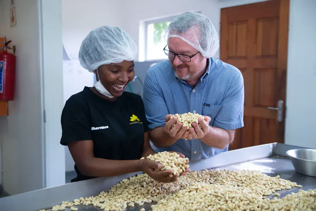 A woman and a man holding and smiling at a pile of light-colored legumes on a table in a processing facility.