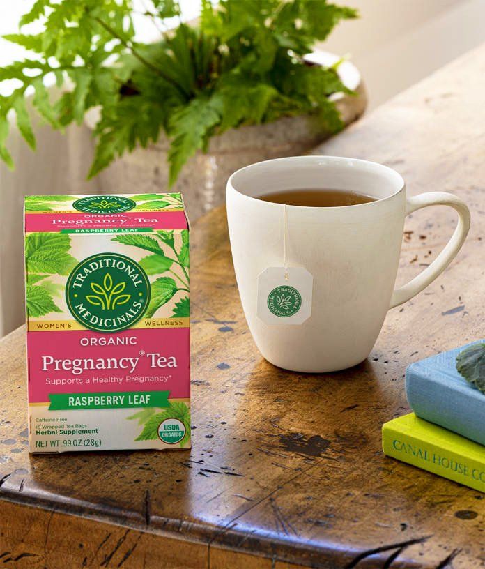 Box of Traditional Medicinals Organic Pregnancy Tea next to a mug of tea on a wooden table, with greenery in the background.
