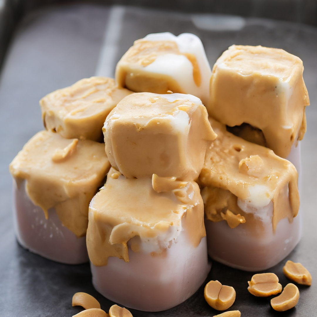 Pile of square, white-chocolate coated candies drizzled with peanut butter, with peanuts scattered around them.