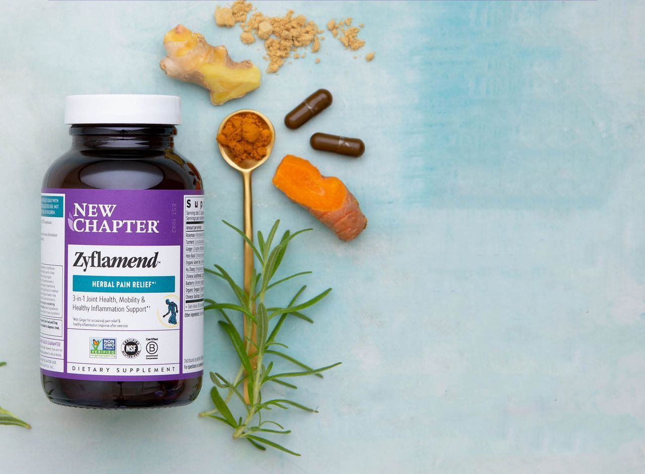 A bottle of New Chapter Zyflamend capsules next to turmeric, ginger, and a sprig of rosemary on a blue background.