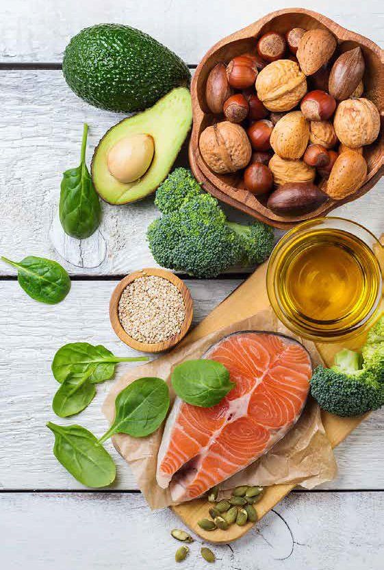 Assortment of healthy fats: avocado, nuts, salmon, broccoli, seeds, and oil, on a light wooden surface.