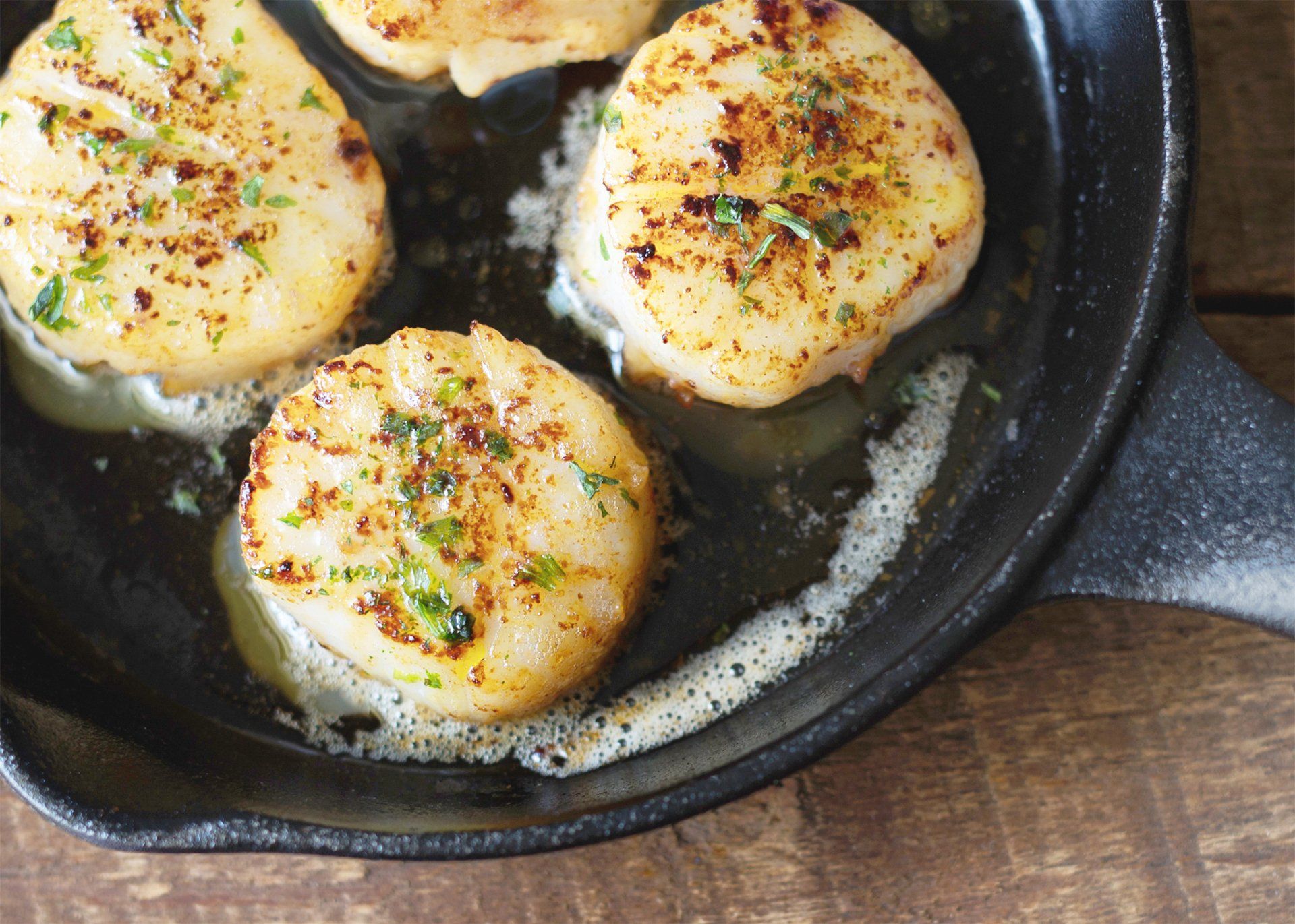 Seared scallops in a cast iron skillet, browned and garnished with herbs, cooking in melted butter.