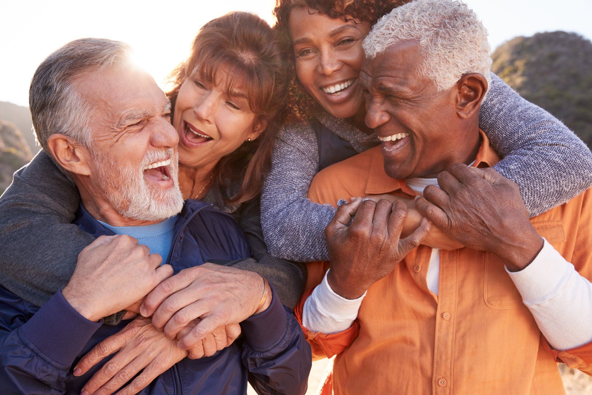 Four diverse people laughing and hugging outdoors. Two women hug two men. Sunny, outdoor setting.