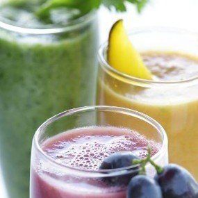 Three colorful smoothies in glasses, one green, one yellow, and one purple, with grapes in front.